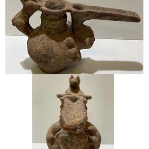 May include: A brown, ancient pottery vessel with a unique design. The vessel features a rounded base, a cylindrical neck, and a long, flat spout. The piece is adorned with animal figures, including a cat-like creature on top.