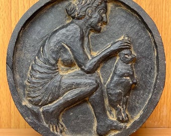May include: A dark gray, circular relief sculpture depicting a person seated and interacting with a cat. The relief is carved with detailed lines and textures, set against a plain background. The sculpture is likely made of stone or a similar material.