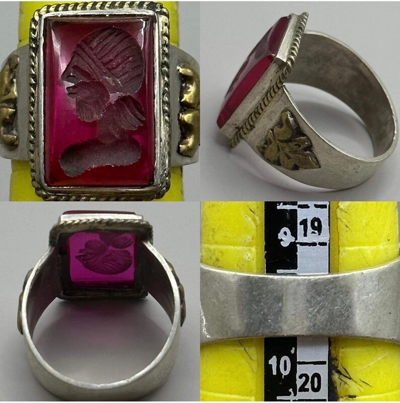 K&ouml;nnte beinhalten: Ein silberner Ring mit einem rechteckigen, tiefroten Edelstein mit einem geschnitzten Profil. Der Ring hat goldfarbene Akzente und einen geflochtenen Rand um den Stein. Die Ringgr&ouml;&szlig;e betr&auml;gt etwa 19 mm.