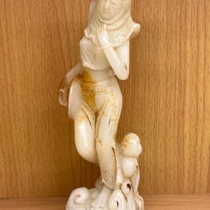 Puede incluir: Una estatuilla tallada, de color blanquecino, de una mujer con un tocado drapeado y ropas fluidas. La figura se alza sobre una base en forma de nube, con una pequeña figura a sus pies. La estatua presenta zonas descoloridas.