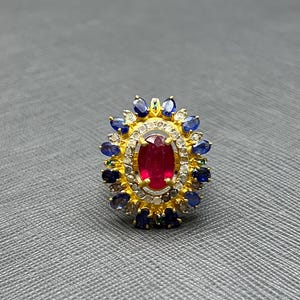 May include: A gold ring featuring a large oval ruby center stone, surrounded by small diamonds and blue sapphires. The ring has a detailed, ornate design with a halo of smaller gemstones. The ring is set on a gray surface.