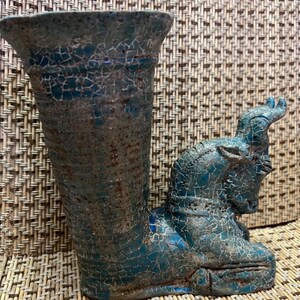Op de afbeelding: Handgemaakte keramische vaas met een turquoise en bruine craquelé glazuur. De vaas heeft een uitlopende bovenkant en een gebeeldhouwde ramfiguur aan de zijkant. De vaas is een uniek decoratief item.