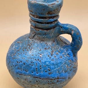 May include: A small, round, blue ceramic jug with a handle. The jug has a textured surface with dark brown speckles and cracks. The top of the jug has a flared opening and a series of horizontal ridges. The handle is curved and attached to the side of the jug.