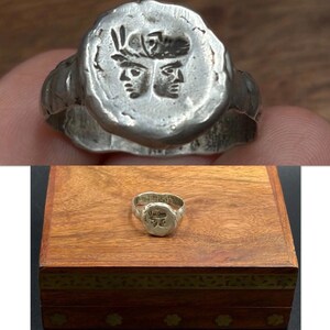 May include: A silver-toned ring featuring a circular face with an embossed design of two faces looking in opposite directions, topped with a feathered headdress. The metal surface has a textured, uneven finish throughout the band and the face.