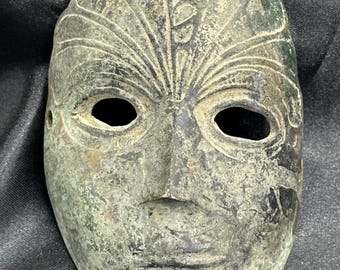 May include: An antique, weathered mask with a human face design. The mask is a muted green-brown color, with open eye sockets and a detailed forehead design. The mask has a distressed appearance, suggesting age and use.