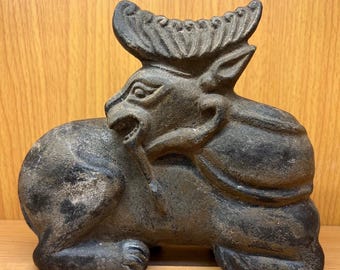 May include: A dark gray, cast iron sculpture of a mythical creature with horns and a detailed mane. The sculpture is in a reclining position, with intricate details on the face and body. The piece has a weathered appearance.