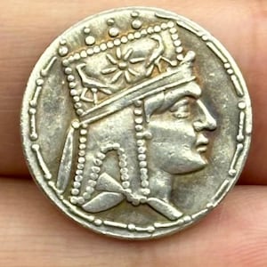 Puede incluir: Una moneda de plata que presenta el perfil de una persona que lleva una corona y un tocado elaborado. El borde de la moneda está detallado con un patrón de puntos. El perfil de la persona mira hacia la derecha, con rasgos faciales detallados.