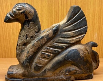 May include: A dark, weathered sculpture of a mythical creature with a bird's head and wings, and the body of a lion. The sculpture is in a seated position, with detailed wing and feather carvings. The color is a mix of black and brown.