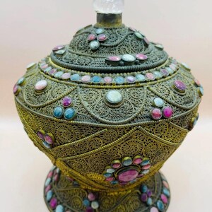 Peut inclure: Un récipient en métal antique orné d'un motif en filigrane. Le récipient est orné de pierres précieuses colorées dans les tons de rose, bleu et vert. Un bouchon transparent en cristal est posé sur le couvercle. Le récipient est probablement à usage décoratif.