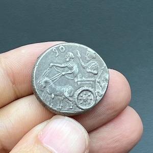 May include: A silver coin depicting a chariot drawn by a horse, with two figures. The coin has a worn look, with details of the chariot, horse, and figures visible. The coin is held in a hand.