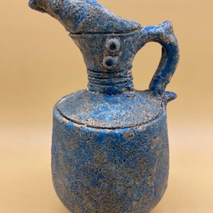 May include: A blue ceramic pitcher with a textured, crackled surface. The pitcher has a spout, handle, and a lid. The design includes two small, round decorative elements near the handle. The overall aesthetic suggests an antique or handcrafted item.