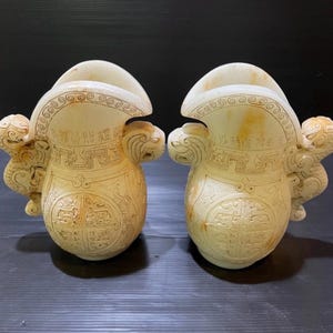 May include: Two off-white, carved stone vessels with intricate designs. Each vessel features a flared top, a rounded body, and detailed relief carvings of mythical creatures and patterns. The vessels are displayed against a dark background.