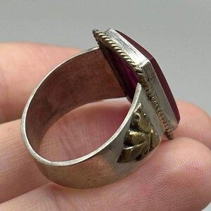 K&ouml;nnte beinhalten: Silberring mit einem rechteckigen, tiefroten Edelstein. Der Ring hat ein dekoratives goldfarbenes Blattdesign an der Seite und einen strukturierten Rand um den Edelstein. Der Ring wird von der Seite gezeigt.