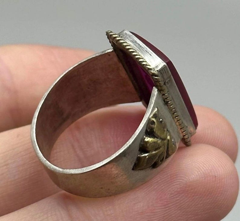 K&ouml;nnte beinhalten: Silberring mit einem rechteckigen, tiefroten Edelstein. Der Ring hat ein dekoratives goldfarbenes Blattdesign an der Seite und einen strukturierten Rand um den Edelstein. Der Ring wird von der Seite gezeigt.