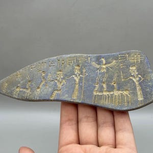 Op de afbeelding: Een blauw, oud artefact met geëtste figuren en symbolen. Het artefact toont afbeeldingen van mensen, dieren en vogels. De figuren zijn lichtbruin van kleur, in contrast met de blauwe achtergrond. Het artefact wordt in de hand van een persoon gehouden.