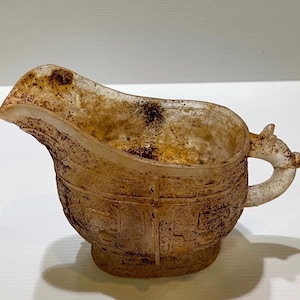 May include: A vintage, off-white, handled vessel with a spout. The vessel has intricate, embossed patterns and a small handle with a decorative element. The piece has a weathered appearance with brown discoloration.