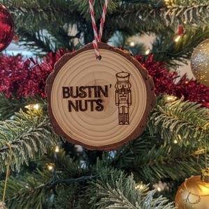 May include: A wooden Christmas tree ornament with the text "BUSTIN' NUTS" and a nutcracker illustration. The ornament is round, made from a cross-section of a tree branch, and hangs from a red and white string. The ornament is displayed on a decorated Christmas tree.