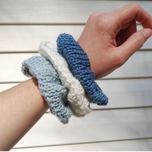 May include: A set of four knitted hair scrunchies in shades of blue and white. The scrunchies are stacked on top of each other, with the darkest blue scrunchie on top and the lightest blue scrunchie on the bottom.