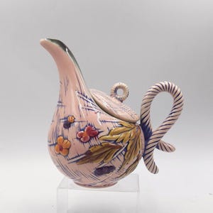 May include: A pink ceramic teapot with a floral design and a twisted handle. The teapot has a lid with a knob on top. The teapot is sitting on a clear acrylic stand.