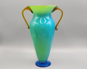 Könnte beinhalten: Eine grüne und blaue Glasvase mit zwei Henkeln. Die Vase hat einen Farbverlauf von Grün zu Blau. Die Henkel sind in einem dunkleren Braunton gehalten.