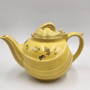 Może przedstawiać: Żółty czajnik z pozłacanym wykończeniem i wzorem liści dębu i żołędzi. Czajnik ma zaokrąglony korpus i zakrzywiony dzióbek.