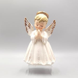 Puede incluir: Una figura de ángel de cerámica con cabello rubio, ojos cerrados y manos juntas en oración. El ángel viste una túnica blanca con detalles de purpurina dorada en el dobladillo y las alas, y presenta un halo dorado sobre la cabeza.