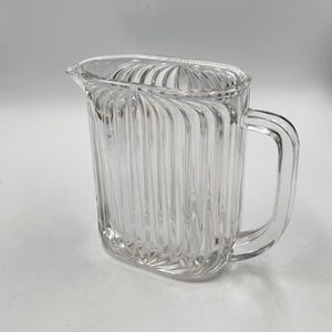 Puede incluir: Una jarra de vidrio transparente con un diseño acanalado. La jarra tiene una forma ovalada redondeada y un asa.