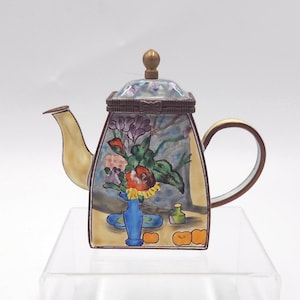 May include: A small, square-shaped metal teapot trinket box featuring a hand-painted floral still life scene on its front panel. The design includes a blue vase, colorful flowers, and orange fruit. It has a curved spout, a handle, and a lid with a knob.