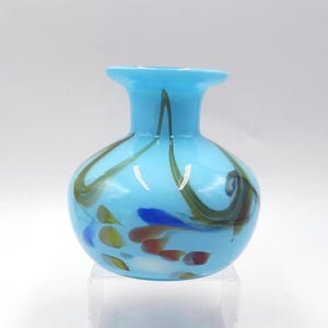 Può includere: Un piccolo vaso di vetro azzurro con corpo arrotondato e bordo svasato. Il vaso è decorato con linee marroni e verdi a spirale e macchie colorate blu, gialle e rosse. Il vaso è probabilmente utilizzato per scopi decorativi.