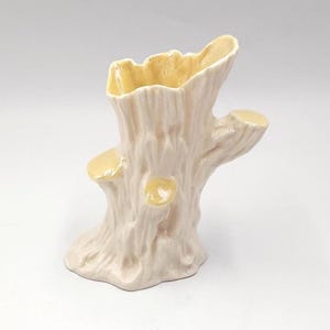 Puede incluir: Un jarrón de cerámica con forma de tocón de árbol. El jarrón es de color blanquecino con una superficie texturizada, y el interior y las ramas cortadas son de color amarillo pálido. El jarrón tiene un diseño único y orgánico.