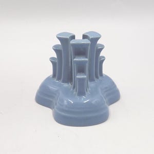 May include: A light blue ceramic candle holder with multiple vertical supports. The candle holder has a rounded base and a series of cylindrical columns of varying heights. The design is symmetrical and the color is a soft, muted blue.