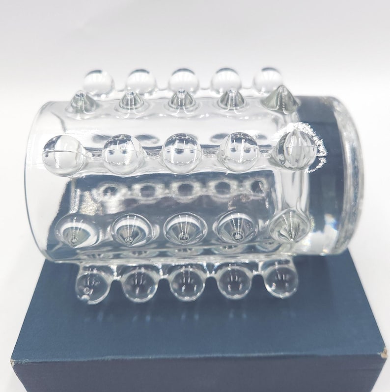 May include: A clear glass cylinder featuring a textured surface with rows of raised, rounded glass studs. The cylindrical object is positioned horizontally, displaying a repeating pattern of spherical protrusions across its entire exterior surface.