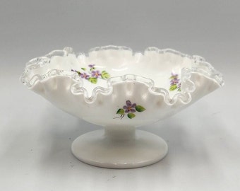 Puede incluir: Un tazón de pedestal de vidrio de leche blanco con un borde rizado con borde plateado. El tazón está decorado con flores moradas pintadas a mano.