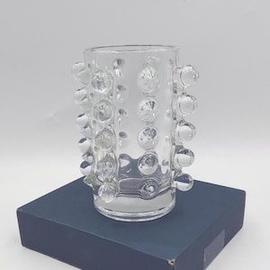 May include: A clear glass vase featuring a cylindrical body adorned with multiple rows of raised, spherical glass bubbles. The vase has a smooth, flat base and an open top, showcasing a transparent design that reflects light across its textured surface.