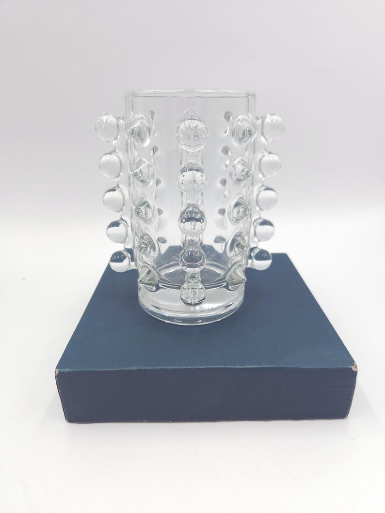 May include: A clear glass vase featuring a cylindrical body adorned with vertical rows of raised, spherical glass studs. The vase has a smooth, open top rim and a flat base, showcasing a transparent design that catches light through its textured surface.