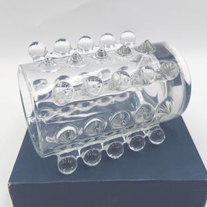May include: A clear glass cuff bracelet featuring a cylindrical shape with a textured surface. The exterior is adorned with multiple rows of raised, rounded glass spheres and pointed glass studs arranged in a repeating pattern around the band.