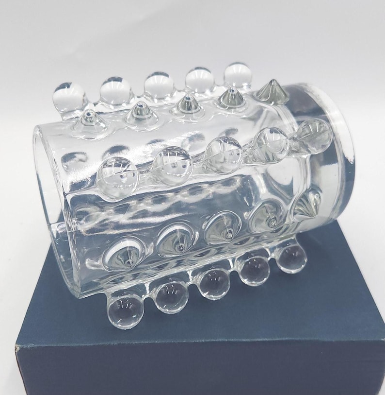 May include: A clear glass cuff bracelet featuring a cylindrical shape with a textured surface. The exterior is adorned with multiple rows of raised, rounded glass spheres and pointed glass studs arranged in a repeating pattern around the band.