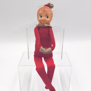 May include: A vintage-style elf doll with a peach-colored face, blue eyes, and a small smile. The doll wears a solid red felt suit with tan cuffs and a matching red pointed hat topped with a small metallic bell. The figure is posed in a seated position.