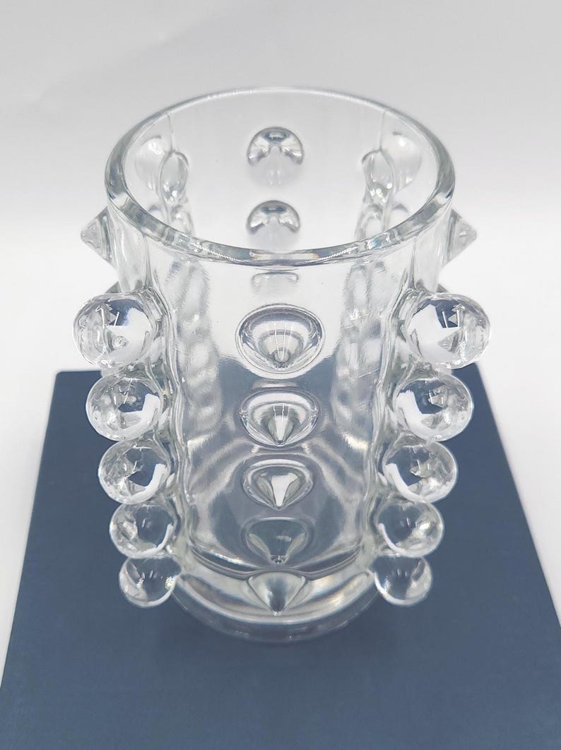 May include: A clear glass cylindrical vase featuring a textured surface with multiple protruding spherical glass bubbles arranged in vertical rows around the exterior. The glass is transparent and smooth, with a wide circular opening at the top.