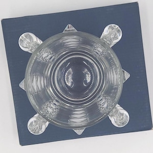 May include: A clear glass bowl with a circular rim and a base featuring four rounded, protruding feet. The exterior of the glass is decorated with a series of vertical, textured ridges that extend from the base toward the upper rim of the bowl.