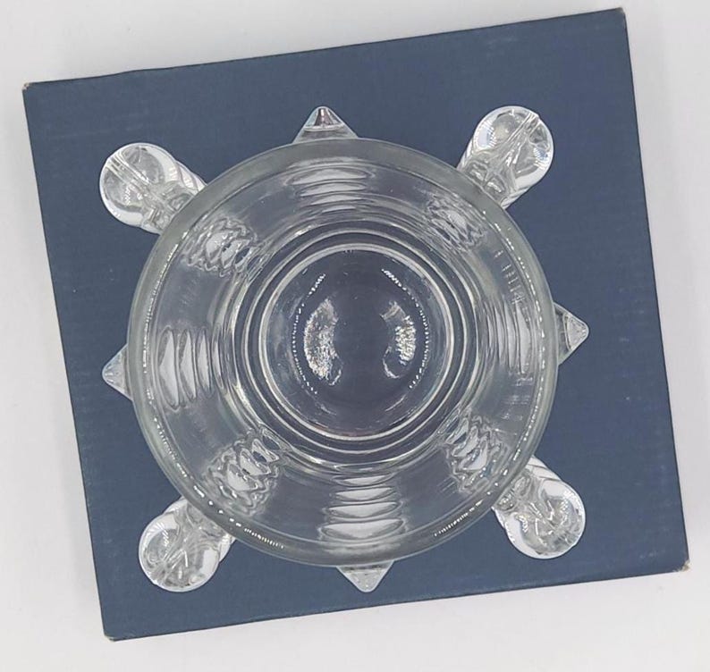 May include: A clear glass bowl with a circular rim and a base featuring four rounded, protruding feet. The exterior of the glass is decorated with a series of vertical, textured ridges that extend from the base toward the upper rim of the bowl.