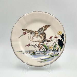 May include: Cream-colored ceramic plate with a bird illustration. The bird has brown and gray wings, a red head, and long beak. The plate has a scalloped edge with a silver rim. The bird is standing in a marsh with yellow flowers.