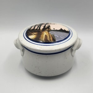 Puede incluir: Jarrón de cerámica blanca con tapa que presenta una escena pintada de un bosque con árboles y un río. El jarrón tiene ribetes azules y blancos y dos asas.