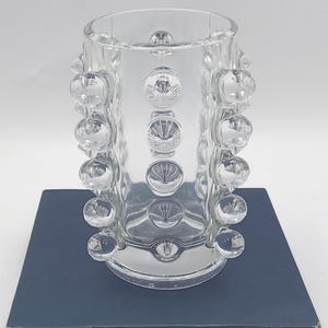 May include: A clear glass cylindrical vase featuring a repeating pattern of raised, spherical glass bubbles arranged in vertical columns around the exterior surface. The vase has a smooth, open top rim and a flat base, showcasing a transparent design.