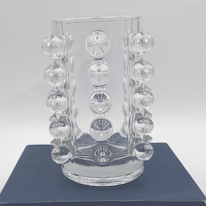 May include: A clear glass vase featuring a cylindrical body adorned with rows of raised, spherical glass bubbles. The design includes vertical columns of these smooth, rounded protrusions that extend along the sides of the transparent vessel.
