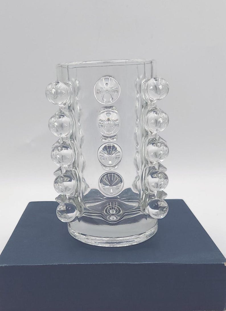 May include: A clear glass vase featuring a cylindrical body adorned with rows of raised, spherical glass bubbles. The design includes vertical columns of these smooth, rounded protrusions that extend along the sides of the transparent vessel.