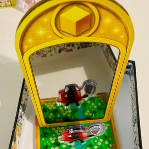 May include: A decorative, yellow-framed mirror with a gold border and a yellow cube design. The mirror reflects a red and silver object on a green surface with white dots. The base has a floral design.