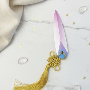 Puede incluir: Un cuchillo decorativo de papel con un diseño geométrico en tonos rosa, morado y azul. Cuenta con una borla dorada y un diseño de nudo. El cuchillo mide aproximadamente 15 cm de largo.