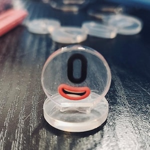 Könnte beinhalten: Ein transparenter Plastikzähler mit der schwarzen Zahl "0" und einem roten Mund. Der Zähler befindet sich auf einer dunklen Holzoberfläche.