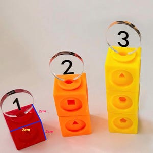 May include: Three colorful stacking blocks with clear plastic number circles on top. The blocks are 2 inches tall and feature different shapes on each side. The numbers are 1, 2, and 3.